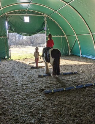 Kind auf einem Tinkerpferd bei einer begleiteten Reitübung in der Reithalle.